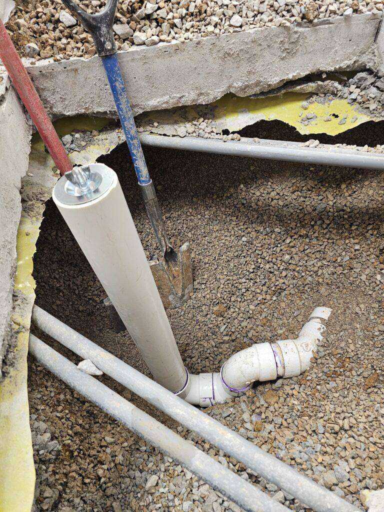 Close-up of exposed PVC pipes and gravel below a broken concrete floor