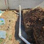 Excavation trench with exposed white plumbing pipe beside a building