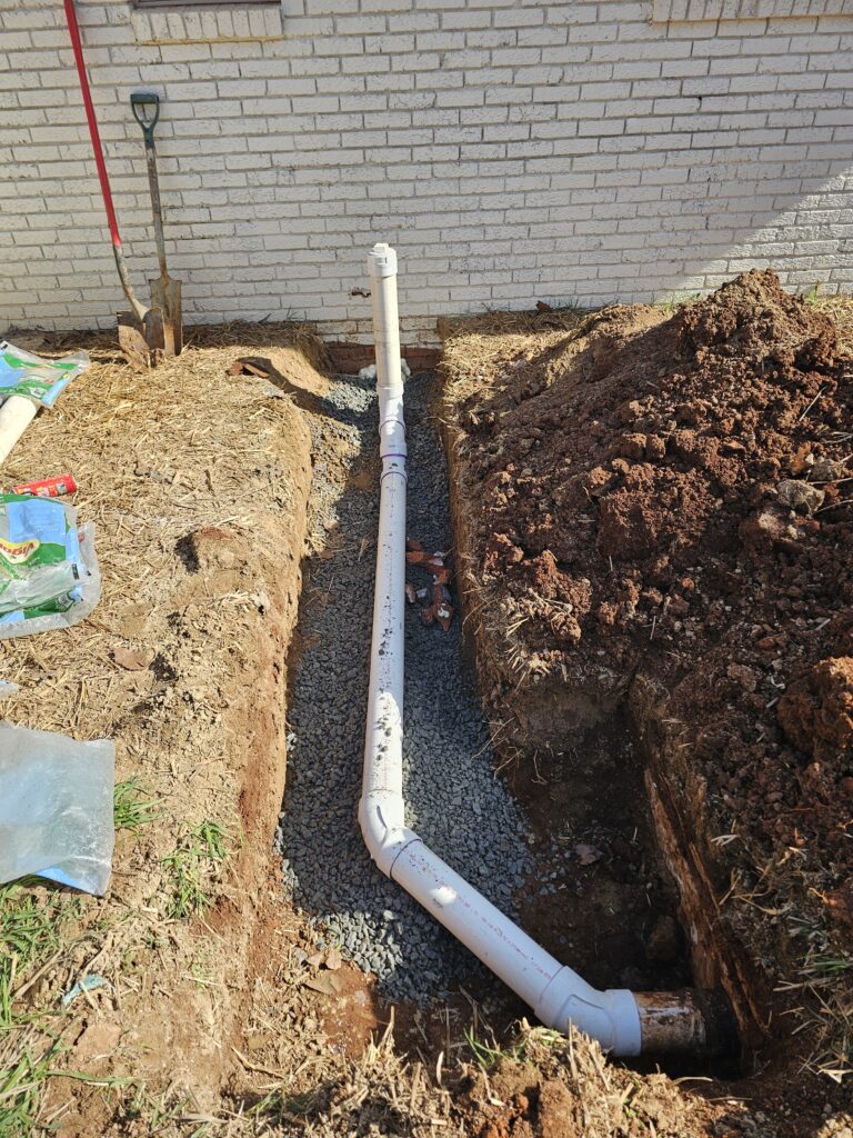 Excavation trench with exposed white plumbing pipe beside a building